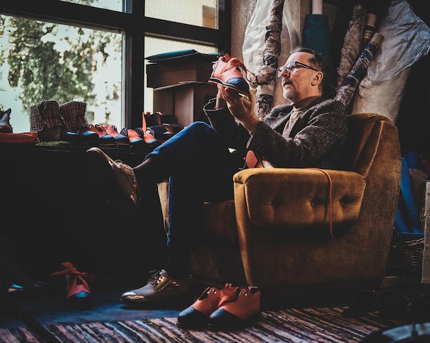 Mature shoe designer is sitting on the armchair at his workshop and holding a shoe from his last collection.