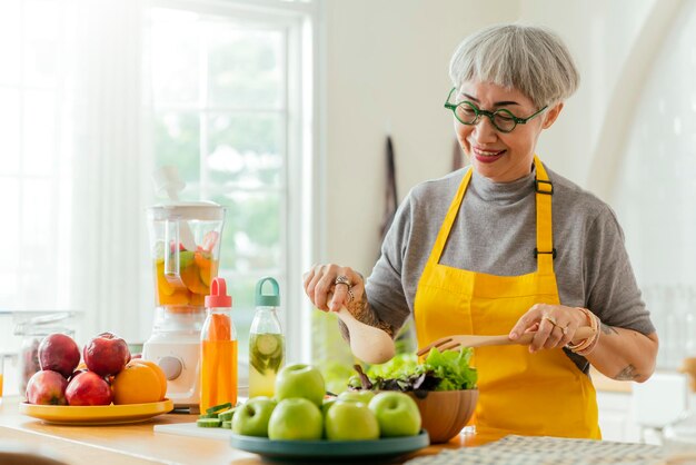 サラダの果物と野菜を食べる成熟した笑顔の入れ墨の女性家で新鮮な緑のフルーツサラダと魅力的な成熟した女性家でリラックスしてキッチンカウンターに立っている年配の女性エプロン