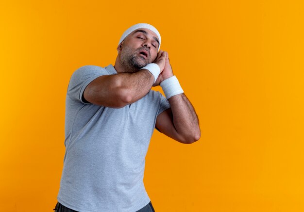 Mature sporty man in headband holding palms together leaning head on palms with closed eyes wants to sleep standing over orange wall