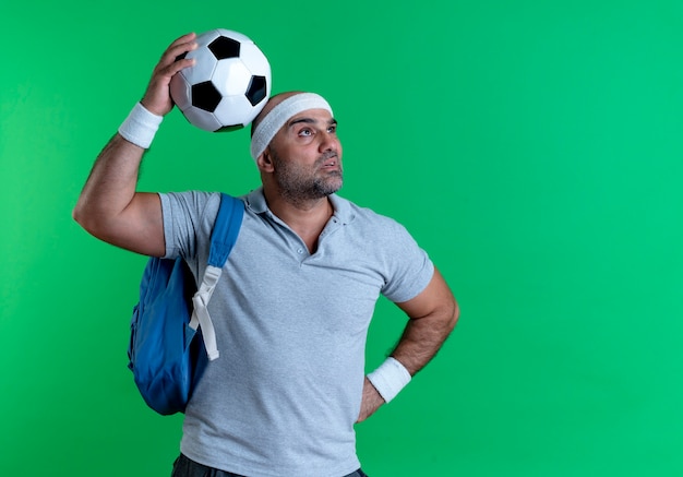 Foto gratuita maturo uomo sportivo in fascia tenendo il pallone da calcio guardando da parte perplesso in piedi sopra la parete verde