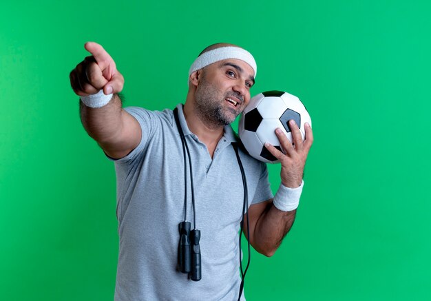 Mature sporty man in headband holding soccer ball looking to the front smiling cheerfully pointing with finger to the front standing over green wall