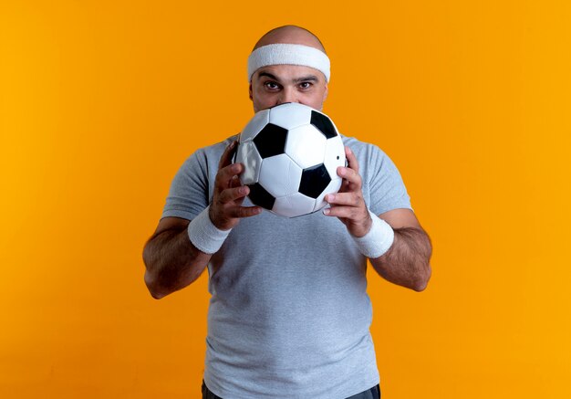 Mature sporty man in headband holding soccer ball looking to the front standing over orange wall