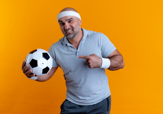 オレンジ色の壁の上に立っている顔に笑顔で正面を向いて指で指しているサッカーボールを保持しているヘッドバンドの成熟したスポーティな男