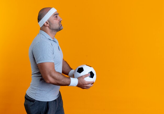 Mature sporty man in headband holding soccer ball standing sideways with serious face over orange wall