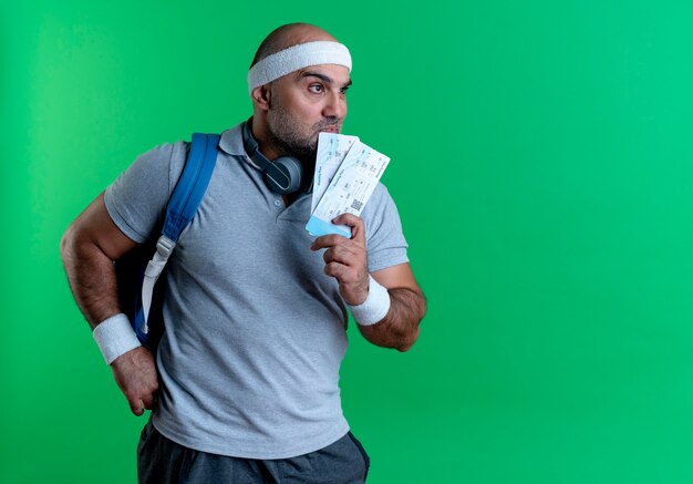 Mature sporty man in headband holding two air tickets looking aside puzzled standing over green wall
