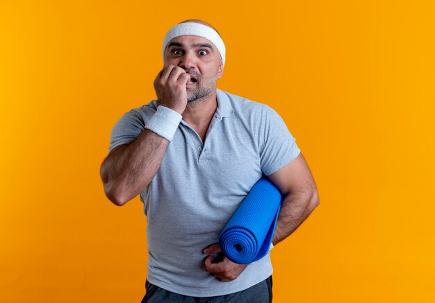 Mature sporty man in headband holding yoga mat stressed and nervous standing over orange wall