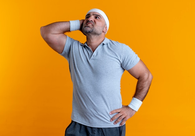 Mature sporty man in headband looking aside with hand on head confused standing over orange wall