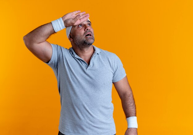 Mature sporty man in headband looking aside with hand over head looking tired and exhausted after workout standing over orange wall