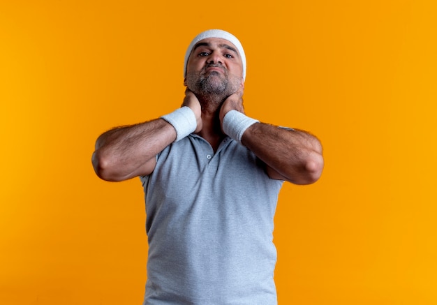 Free photo mature sporty man in headband looking to the front displeased touching his neck feeling discomfort standing over orange wall