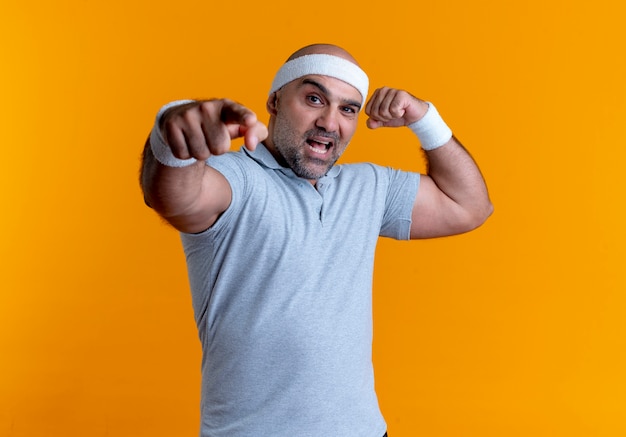 Mature sporty man in headband looking to the front showing biceps pointing with finger to the front smiling standing over orange wall