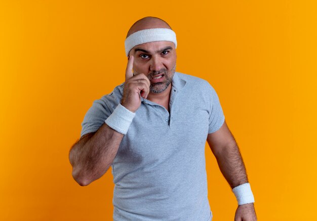 Mature sporty man in headband looking to the front showing index finger warning with serious face standing over orange wall