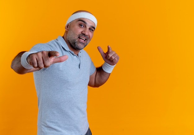 Mature sporty man in headband looking to the front smiling confident pointing with index fingers to the front standing over orange wall