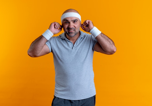 Mature sporty man in headband looking to the front smiling showing his ears standing over orange wall