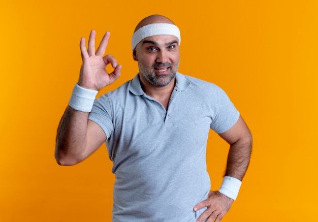 Mature sporty man in headband looking to the front smiling showing ok sign standing over orange wall