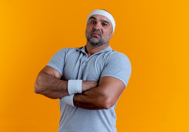 Mature sporty man in headband looking to the front with crossed hands on chest with serious expression standing over orange wall