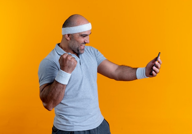 Mature sporty man in headband looking at screen of his mobile phone happy and excited clenching fist standing over orange wall