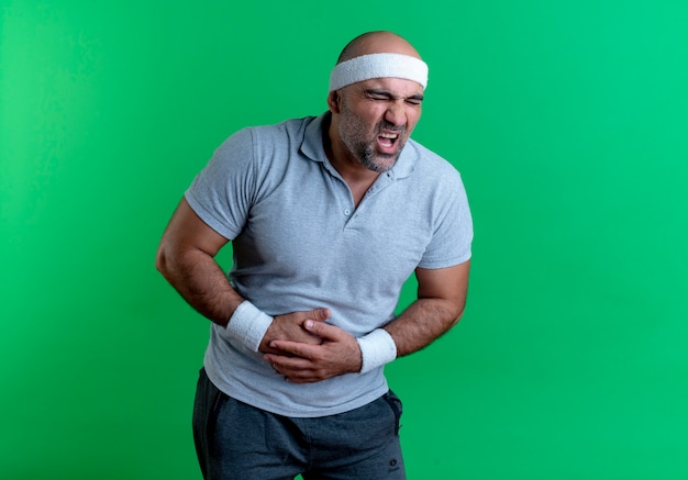 Mature sporty man in headband looking unwell touching his belly suffering from pain standing over green wall