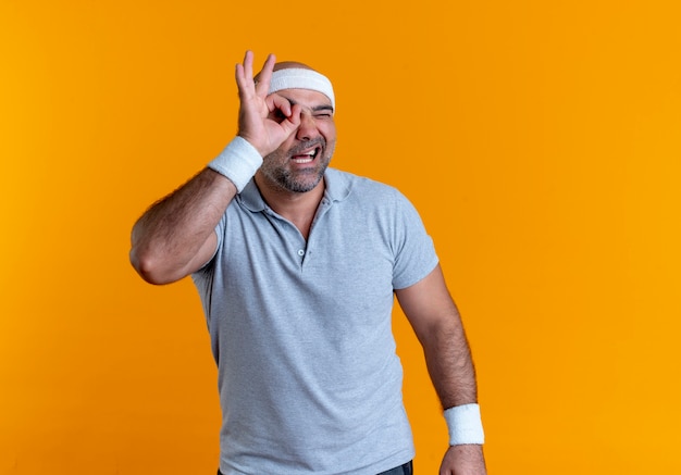 Mature sporty man in headband making ok sign with fingers looking through this sign standing over orange wall
