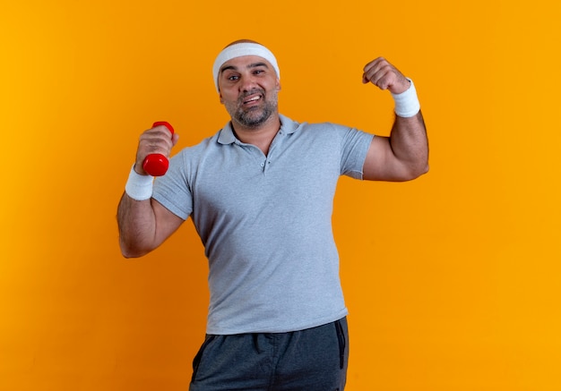 Зрелый спортивный мужчина в повязке на голову, поднимающий руку с гантелями, глядя вперед с уверенным выражением лица, улыбаясь, стоя над оранжевой стеной