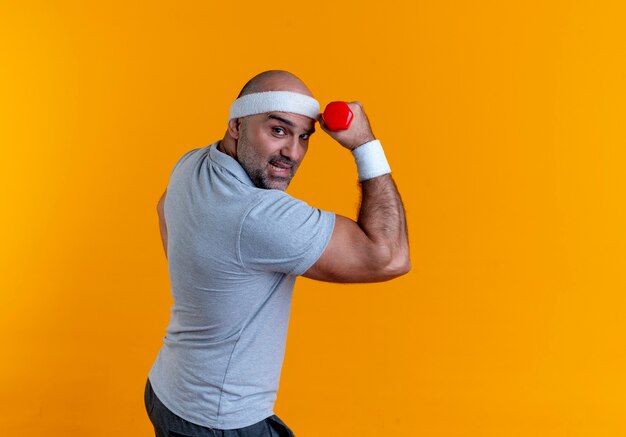 Mature sporty man in headband raising hand with dumbbell looking strained and confident standing over orange wall