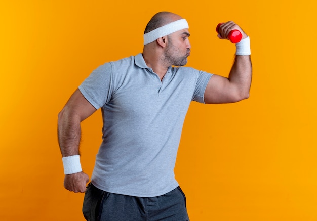 Mature sporty man in headband raising hand with dumbbell showing biceps looking confident standing over orange wall