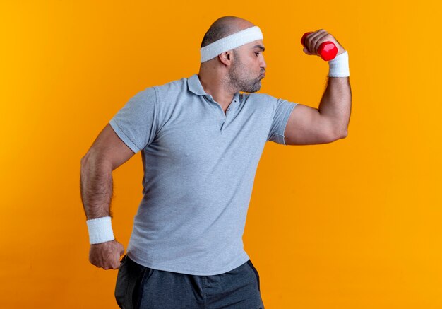 Mature sporty man in headband raising hand with dumbbell showing biceps looking confident standing over orange wall