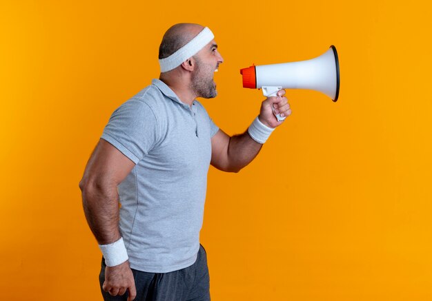Зрелый спортивный мужчина в повязке на голову кричит в мегафон, стоя над оранжевой стеной