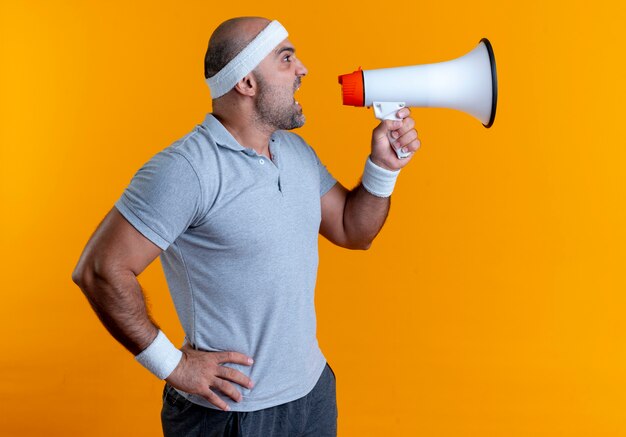 Mature sporty man in headband shouting to megaphone with aggressive expression standing over orange wall