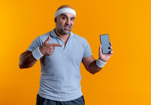 Зрелый спортивный мужчина в повязке на голову показывает смартфон, указывая пальцем на него, выглядит уверенно, стоя над оранжевой стеной