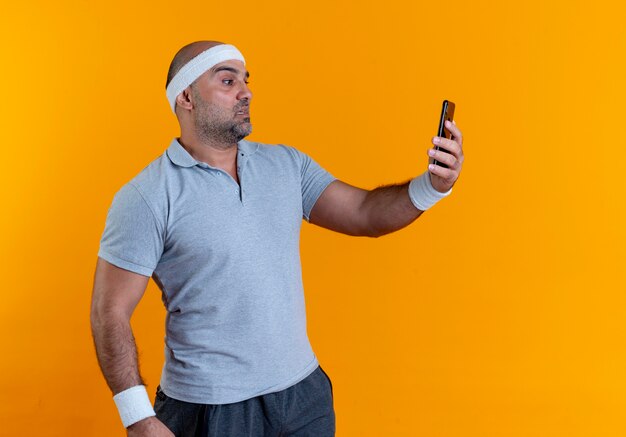 Зрелый спортивный мужчина в повязке на голову, делающий селфи на своем смартфоне, выглядит смущенным, стоя у оранжевой стены