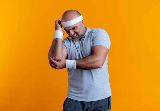 Free photo mature sporty man in headband touching elbow looking unwell feeling pain standing over orange wall