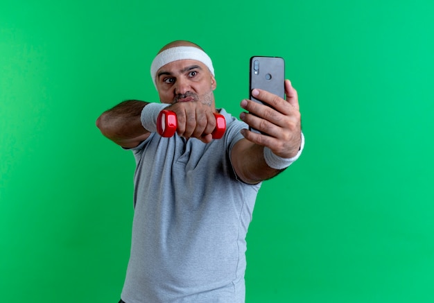 Зрелый спортивный мужчина в повязке на голову с полотенцем на шее, поднимающий руку с гантелью, делает селфи на своем смартфоне и смотрит с серьезным лицом, стоящим над зеленой стеной