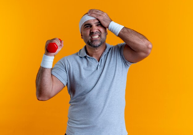 Mature sporty man in headband working out with dumbbell looking confused standing over orange wall