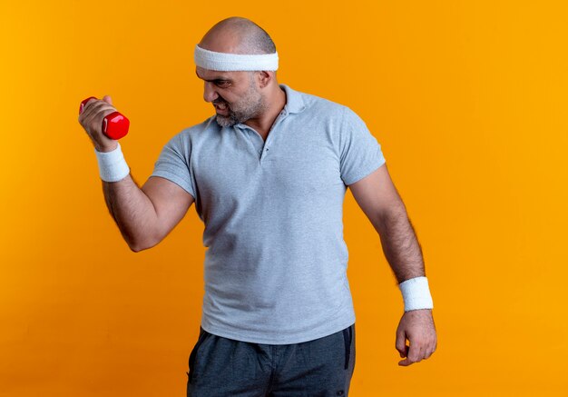 Mature sporty man in headband working out with dumbbell looking strained and confident standing over orange wall