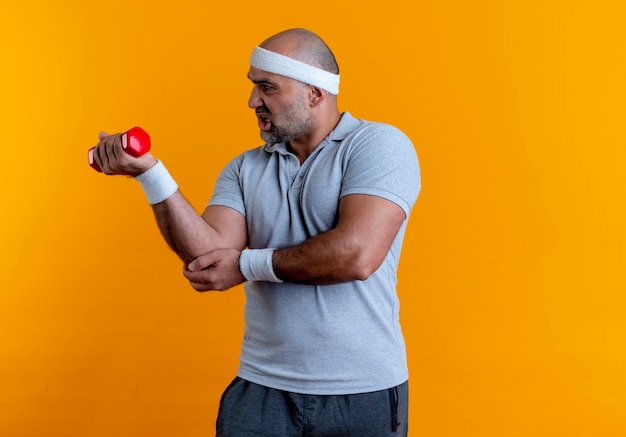 Free photo mature sporty man in headband working out with dumbbell looking tired and exhausted standing over orange wall