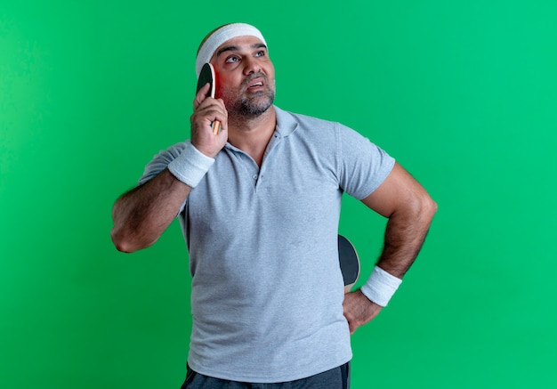Зрелый спортивный мужчина в повязке на голову, держащий две ракетки для настольного тенниса, озадаченно смотрит в сторону, стоя у зеленой стены