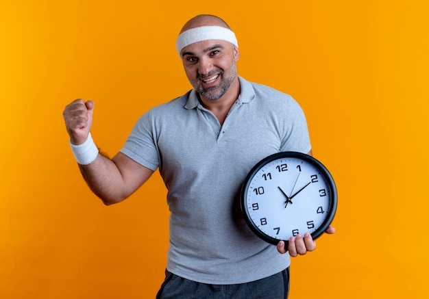 Зрелый спортивный мужчина в повязке на голову держит настенные часы, сжимая кулак, счастливый и взволнованный, стоя над оранжевой стеной