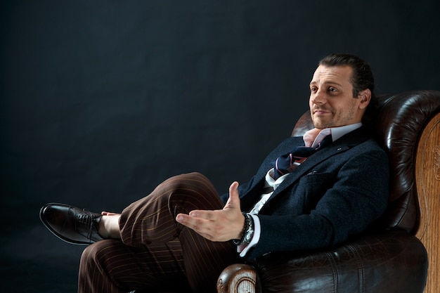 灰色のスタジオでスーツを着た成熟したスタイリッシュな男。肘掛け椅子に座っている実業家