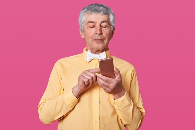 зрелый седой мужчина, одетый в желтую рубашку и белый галстук-бабочку, держит смартфон в руках и печатает сообщение