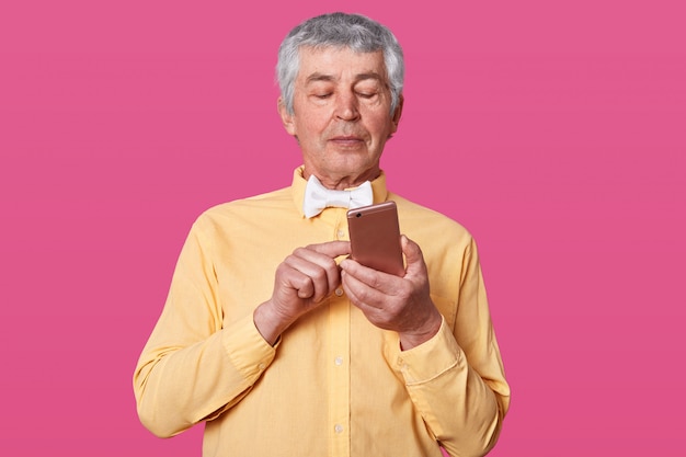 mature white haired man dressed in yellow shirt and white bow tie, holding smartphone in hands and types message