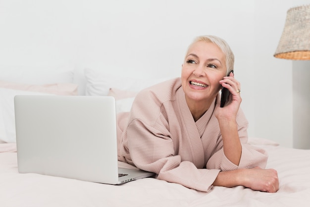 Foto gratuita donna matura in accappatoio che parla sul telefono e che posa con il computer portatile