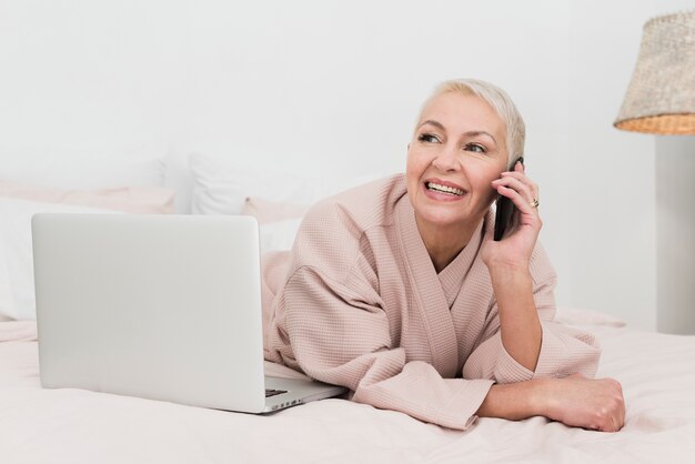 Mature woman in bathrobe talking on phone and posing with laptop