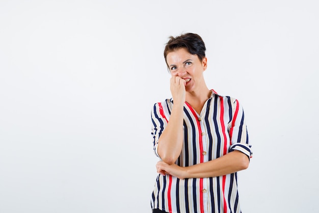 Mature woman biting fingers, holding hand under elbow in striped shirt and looking excited. front view.