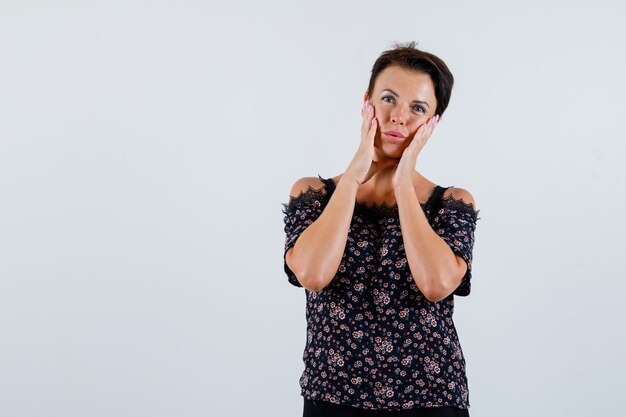 Mature woman in blouse holding hands on cheeks and looking pensive , front view.