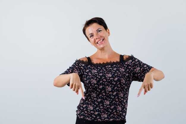 Mature woman in blouse pointing down and looking happy , front view.