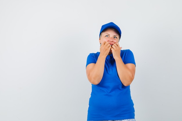 Mature woman in blue t-shirt biting fists emotionally and looking scared.