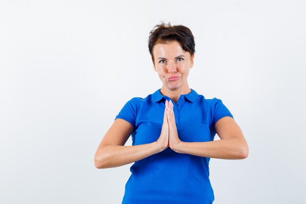 Mature woman in blue t-shirt showing namaste gesture and looking hopeful , front view.