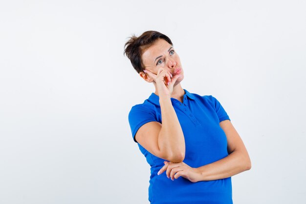 Зрелая женщина в голубой футболке, стоящая в позе мышления и выглядящая разумно, вид спереди.