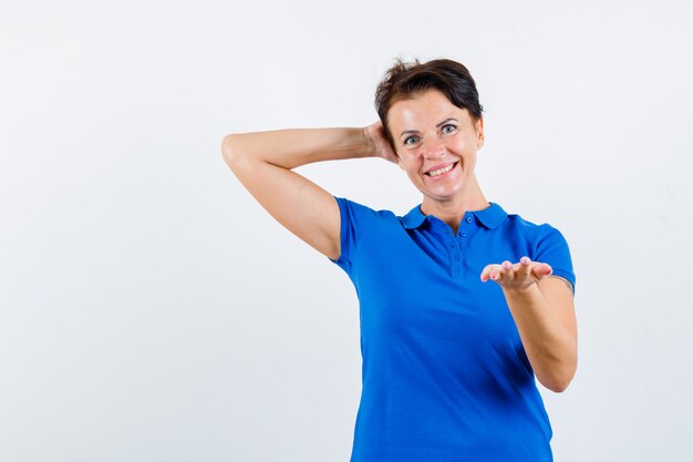 Зрелая женщина в голубой футболке протягивает руку, держа другую руку за голову и выглядит счастливой, вид спереди.