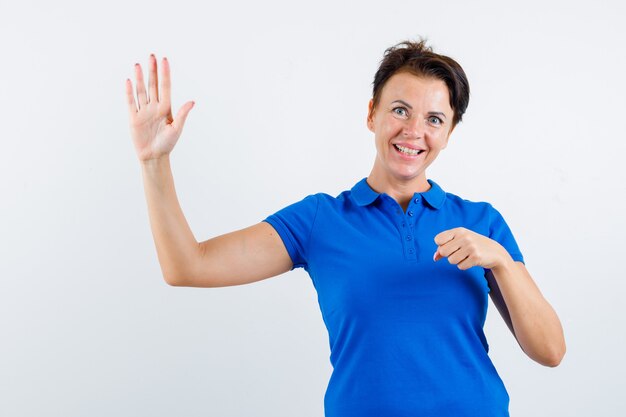 Mature woman in blue t-shirt waving hand while keeping fist clenched and looking merry , front view.